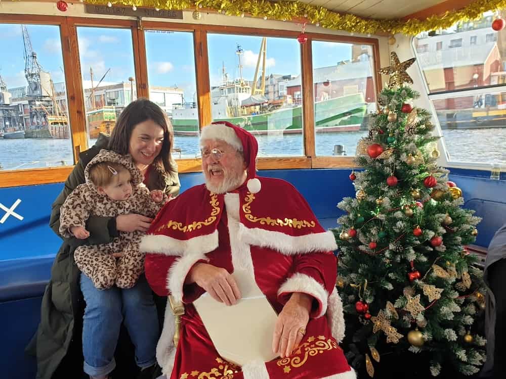 Father Christmas on a Bristol ferry boat smiling at a woman with a toddler on her lap. 