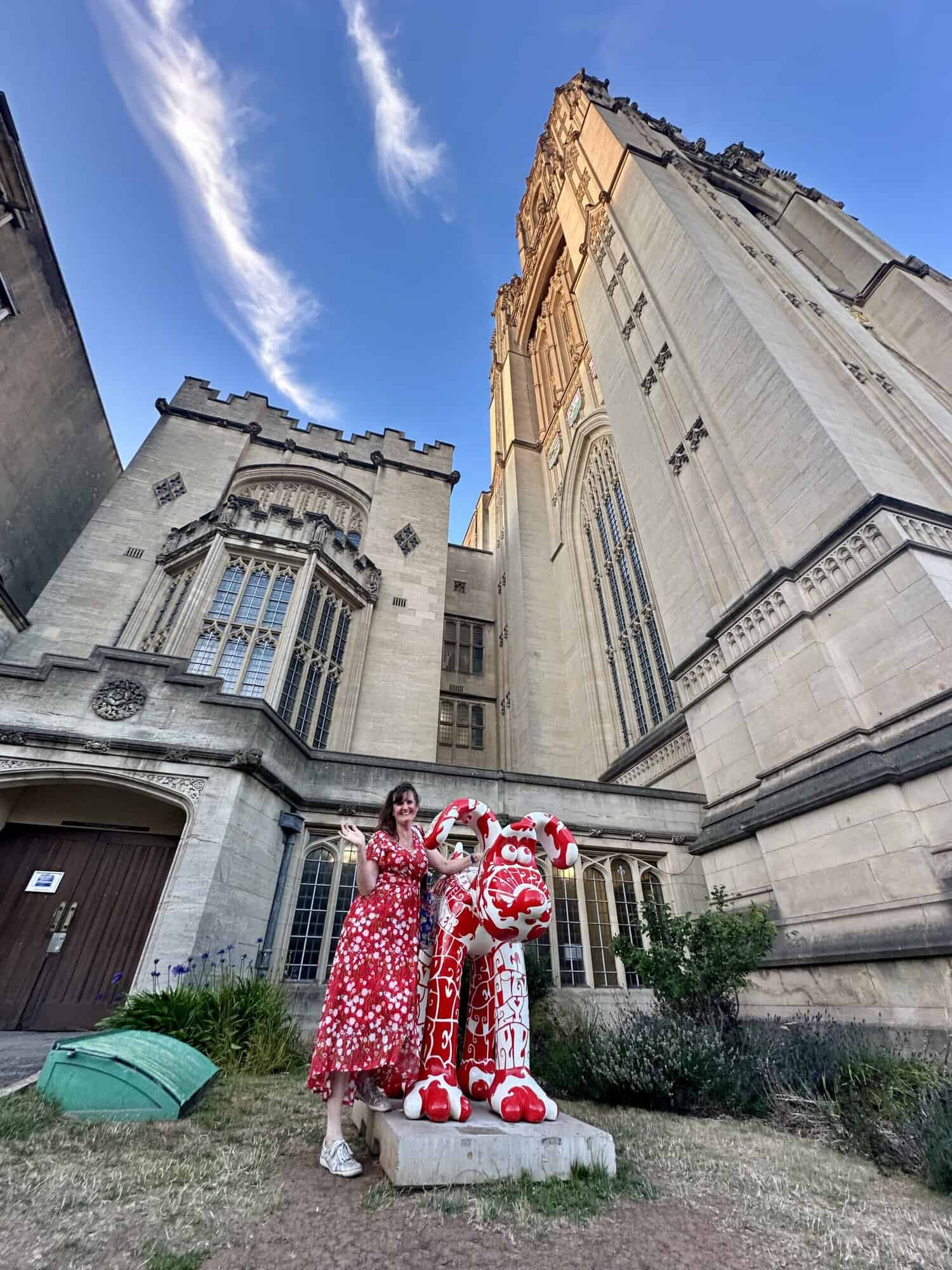 Jane Batt posing with a Gromit Unleashed statue near the top of Park Street with The Wills Building to one side and bright blue sky above. Jane's red and white floaty summer dress matches Gromit the dog's red and white patterned colour-scheme.